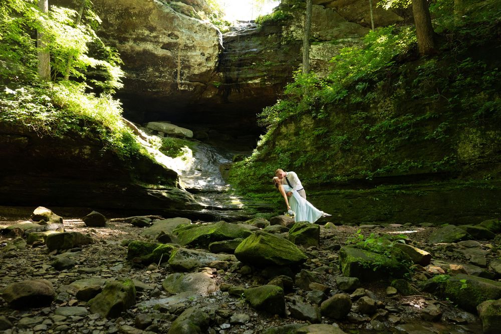 A bride in a white dress poses on mossy rocks beside a waterfall in a lush green canyon.