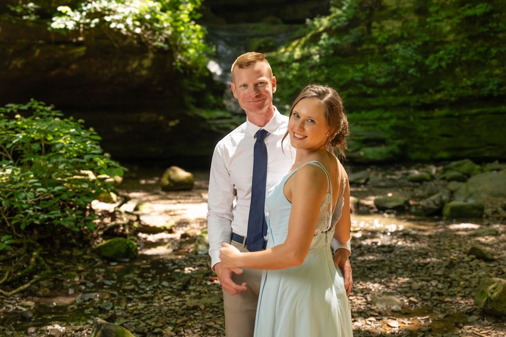 Couple posing outdoors in a lush green forest setting, man in white shirt and blue tie, woman in light blue dress.