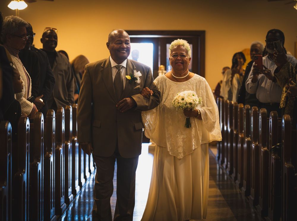 Elderly couple walks down church aisle during wedding ceremony, bride in white gown holding bouquet, groom in suit.