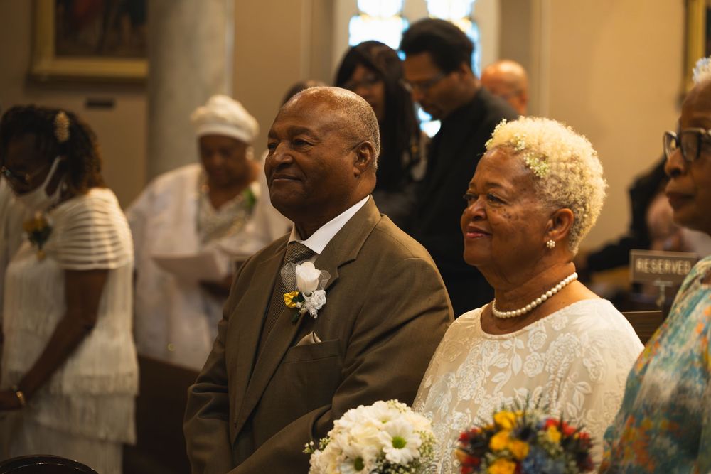 Elderly couple smiling at a wedding ceremony, man in suit with boutonniere, woman in white lace dress with pearl necklace.