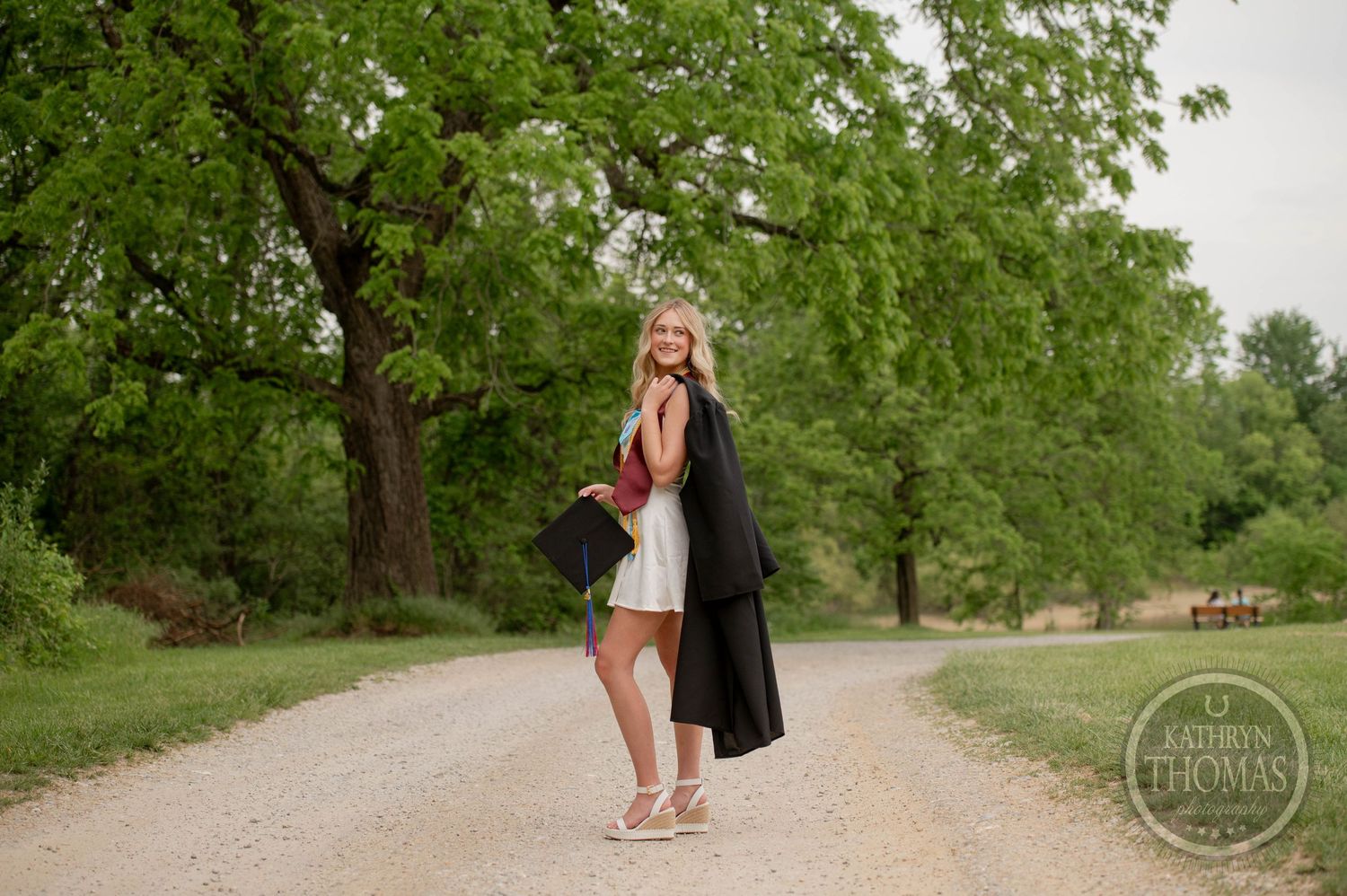 Graduate in black cap and gown stands on gravel path surrounded by green trees during outdoor photo session.