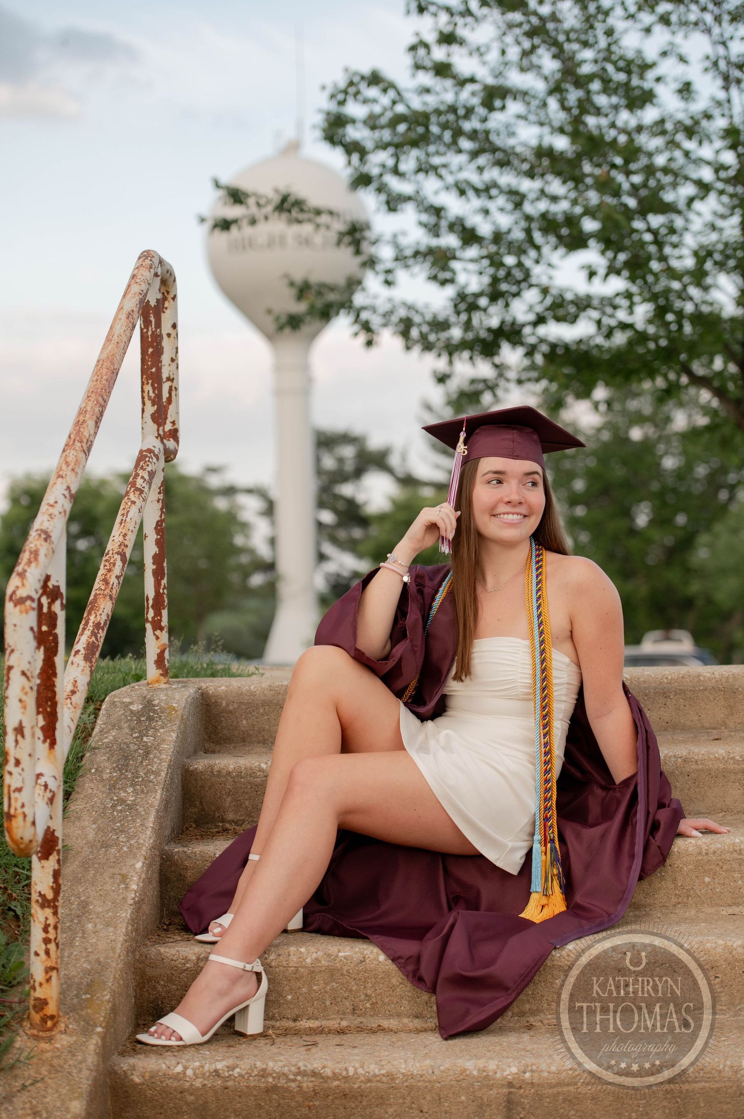 Evening graduation portrait shows graduate in maroon cap and gown seated on concrete steps with water tower in background.