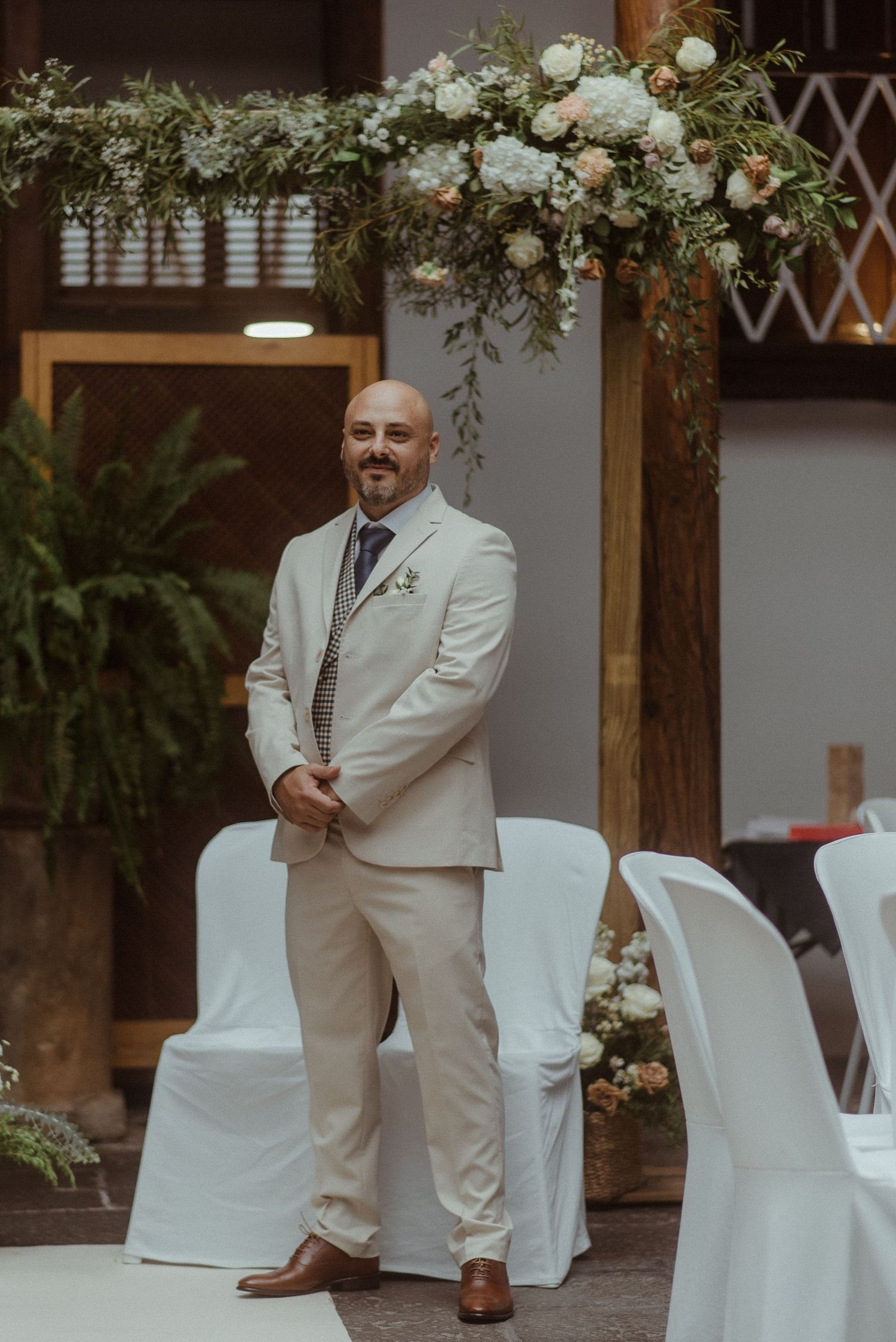 A wedding ceremony setup with a wooden arch decorated with white flowers and greenery, with white covered chairs arranged below.