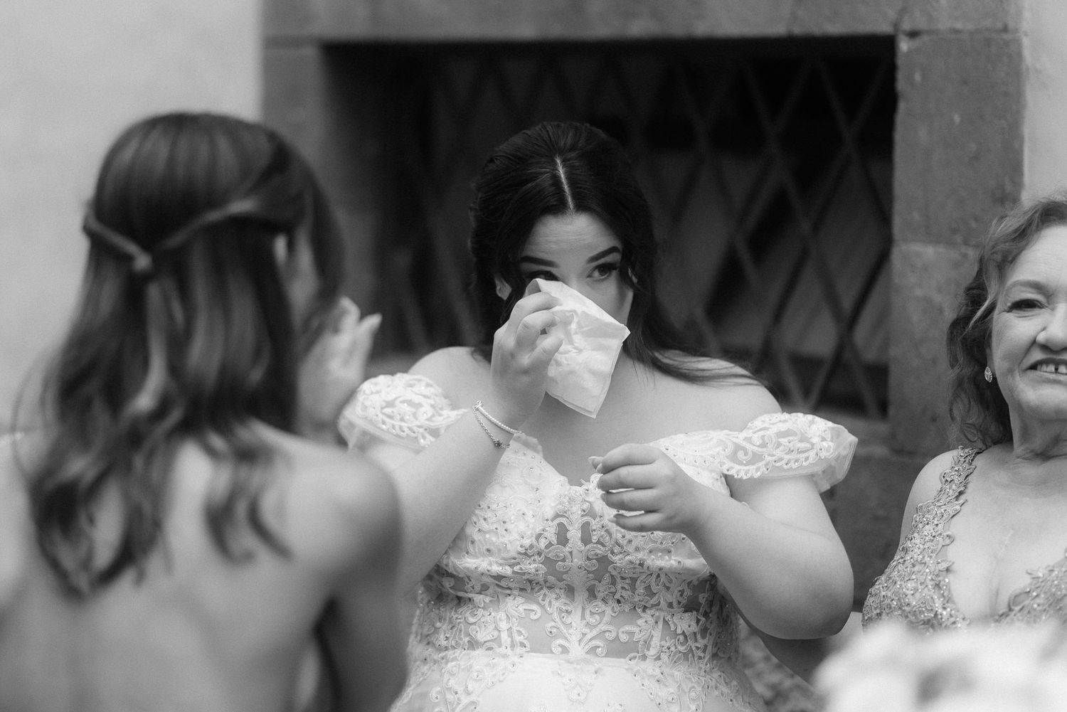 A bride wiping her nose while standing with two other women wearing formal dresses in an emotional black and white photo.