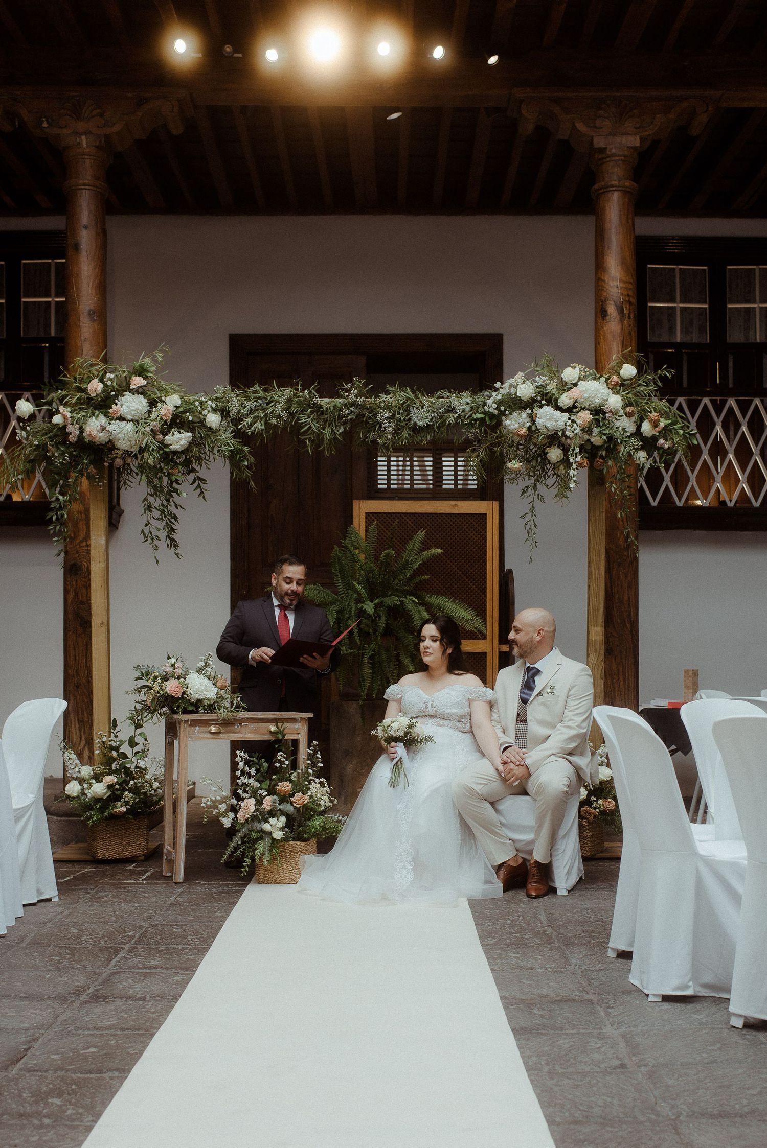 Wedding ceremony setup with white floral arch, covered chairs and a white aisle runner in an elegant indoor venue with columns.