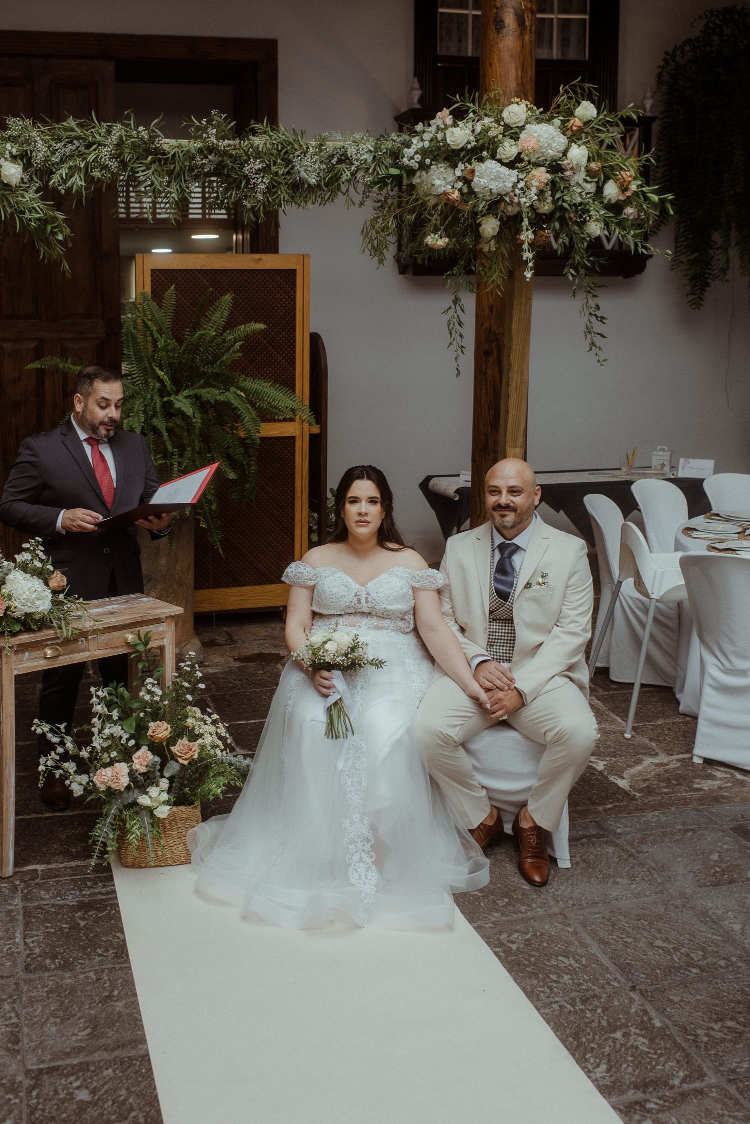 A couple sits during their wedding ceremony with elegant white floral arrangements and an officiant in a red tie.