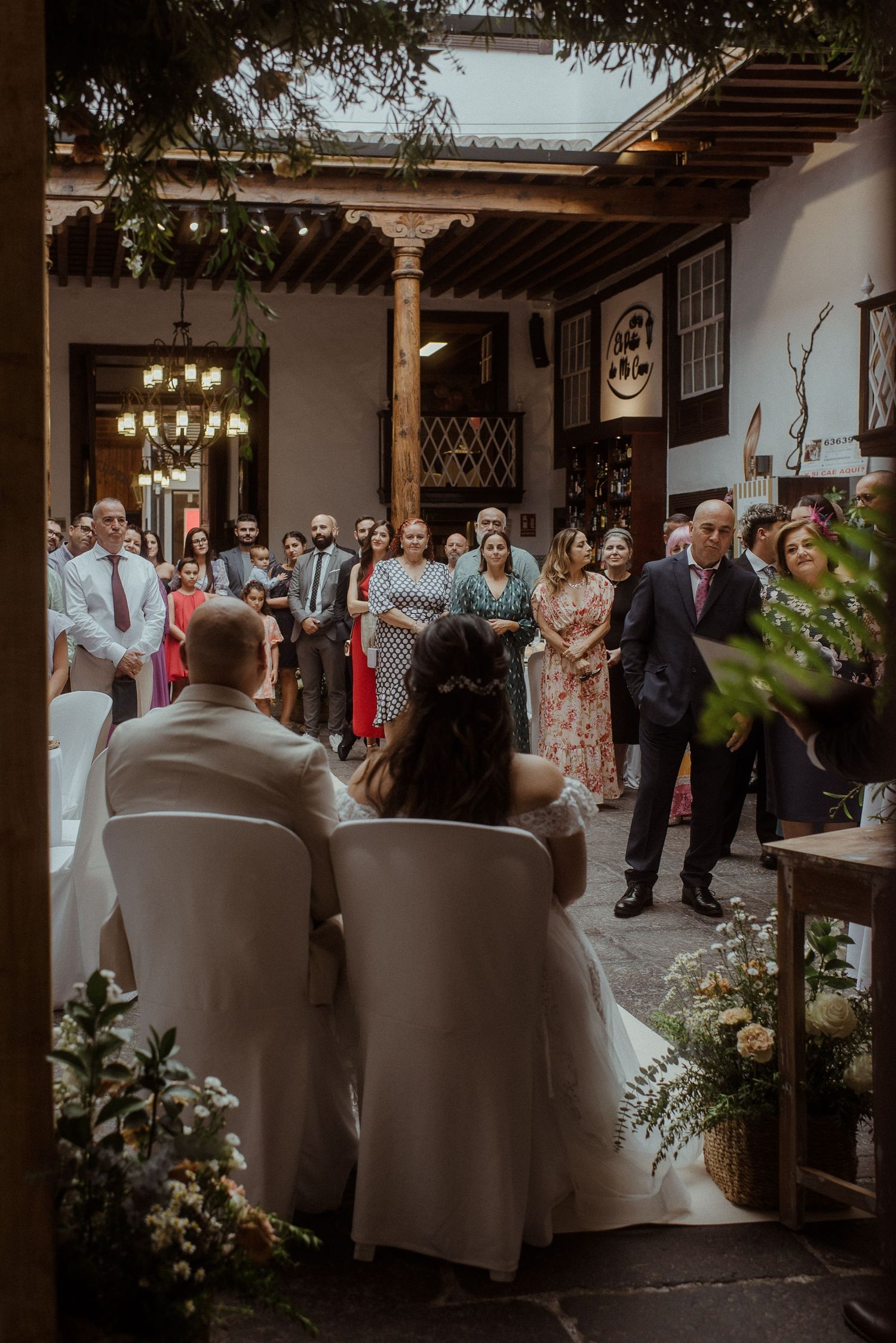 Wedding ceremony guests gather in an elegant courtyard with wooden beams and chandelier lighting during a sunset celebration.