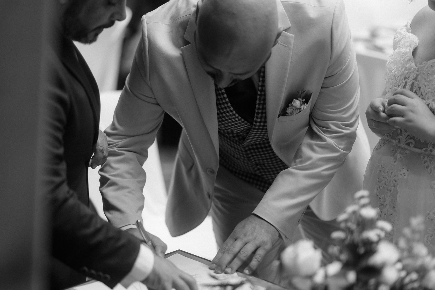 A person in a light colored suit leans over a surface while interacting with others in a black and white photograph.