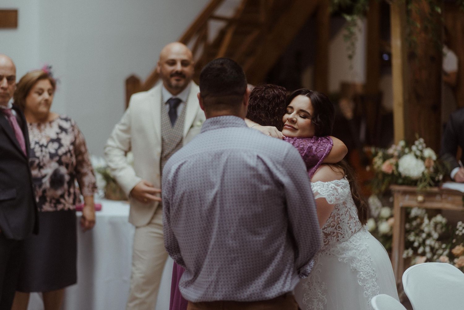 Newlyweds embrace during their wedding reception at a rustic indoor venue with guests looking on.