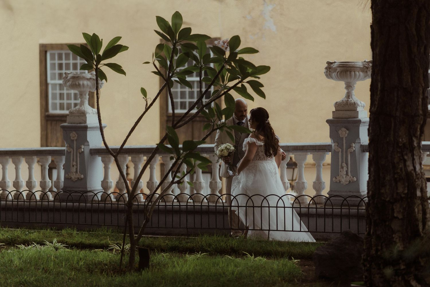 A person in a wedding gown stands on an ornate balcony beside a small tree at dusk.