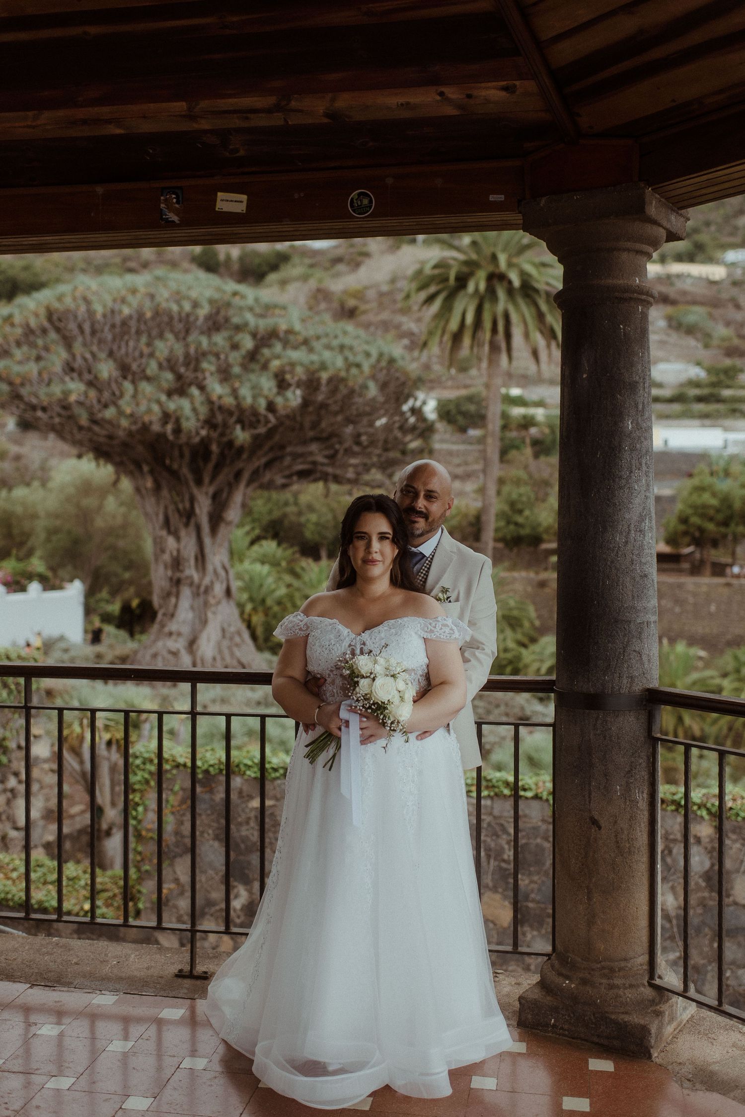 A couple stands beneath a covered patio with stone pillars and wooden beams near a stunning ancient dragon tree.