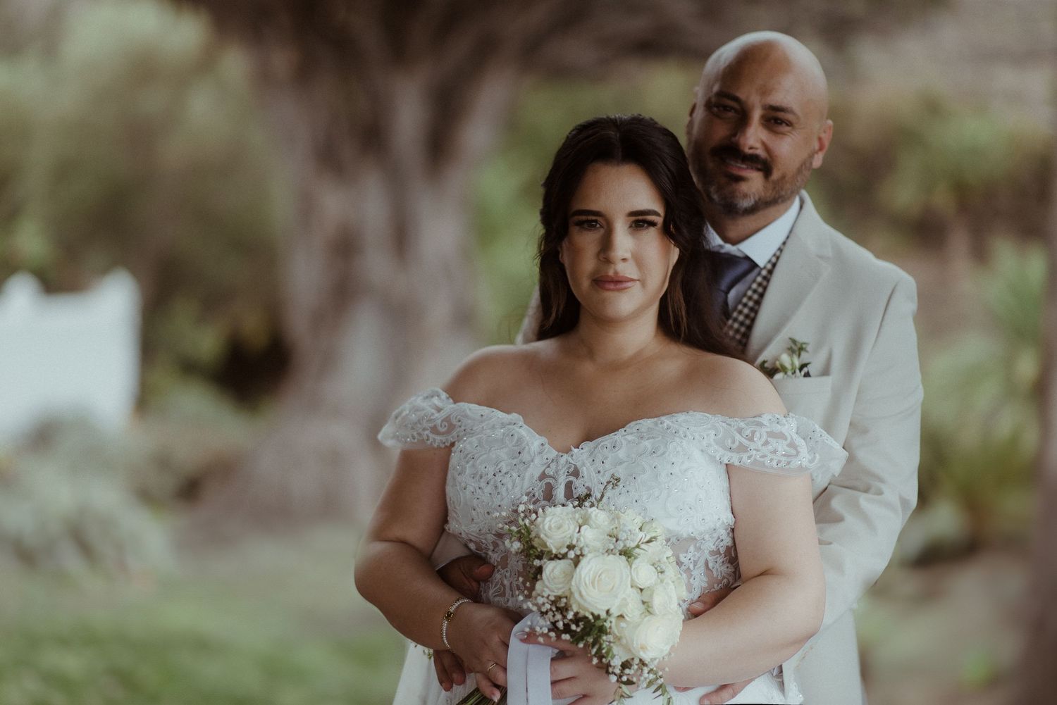 A newlywed couple poses outdoors during their wedding, with the groom in white and bride in lace gown.