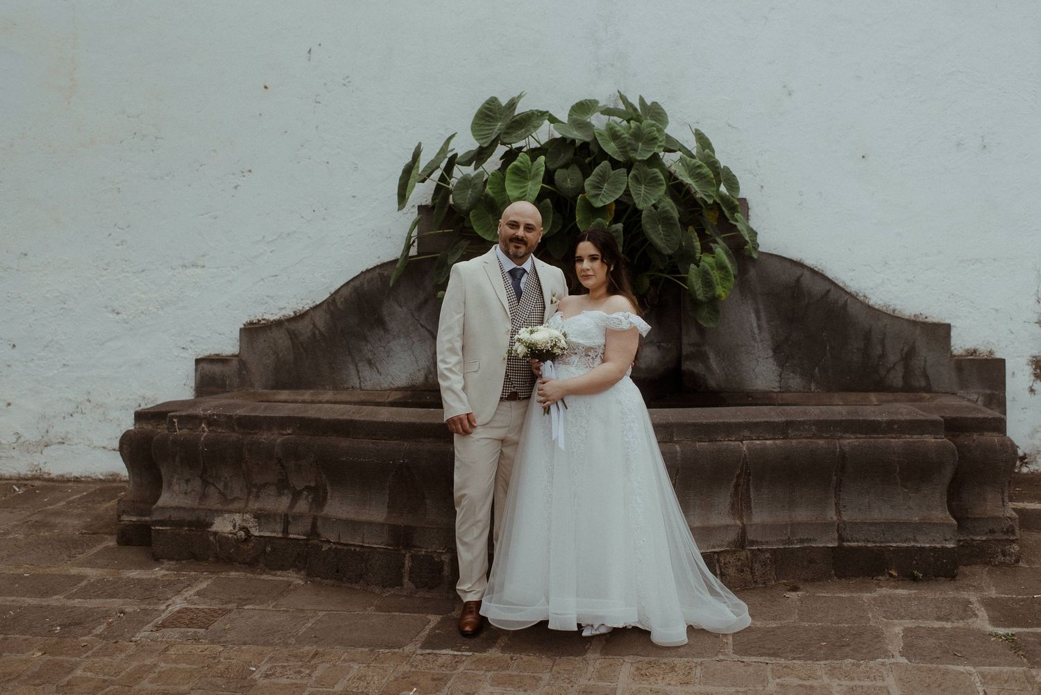 A couple in wedding attire stands in front of an ornate stone bench with greenery at an old architectural setting.