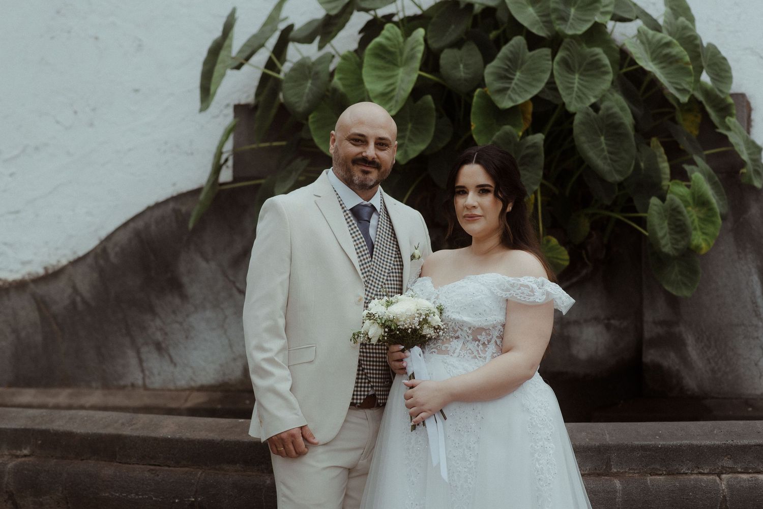A couple in wedding attire poses outdoors against a wall with lush green foliage, the groom in white and bride holding flowers.