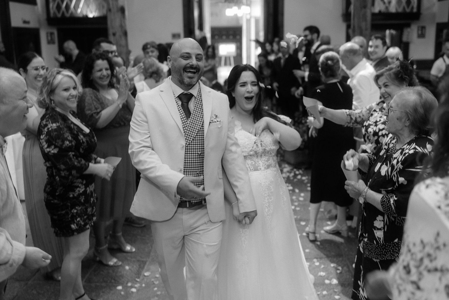 Joyful newlyweds walk down the aisle while wedding guests shower them with flower petals in black and white.