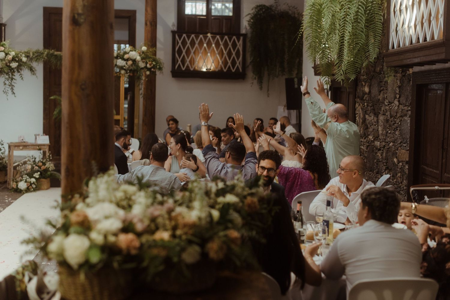 Wedding guests gather at a rustic indoor reception venue decorated with elegant floral arrangements and wooden beams.
