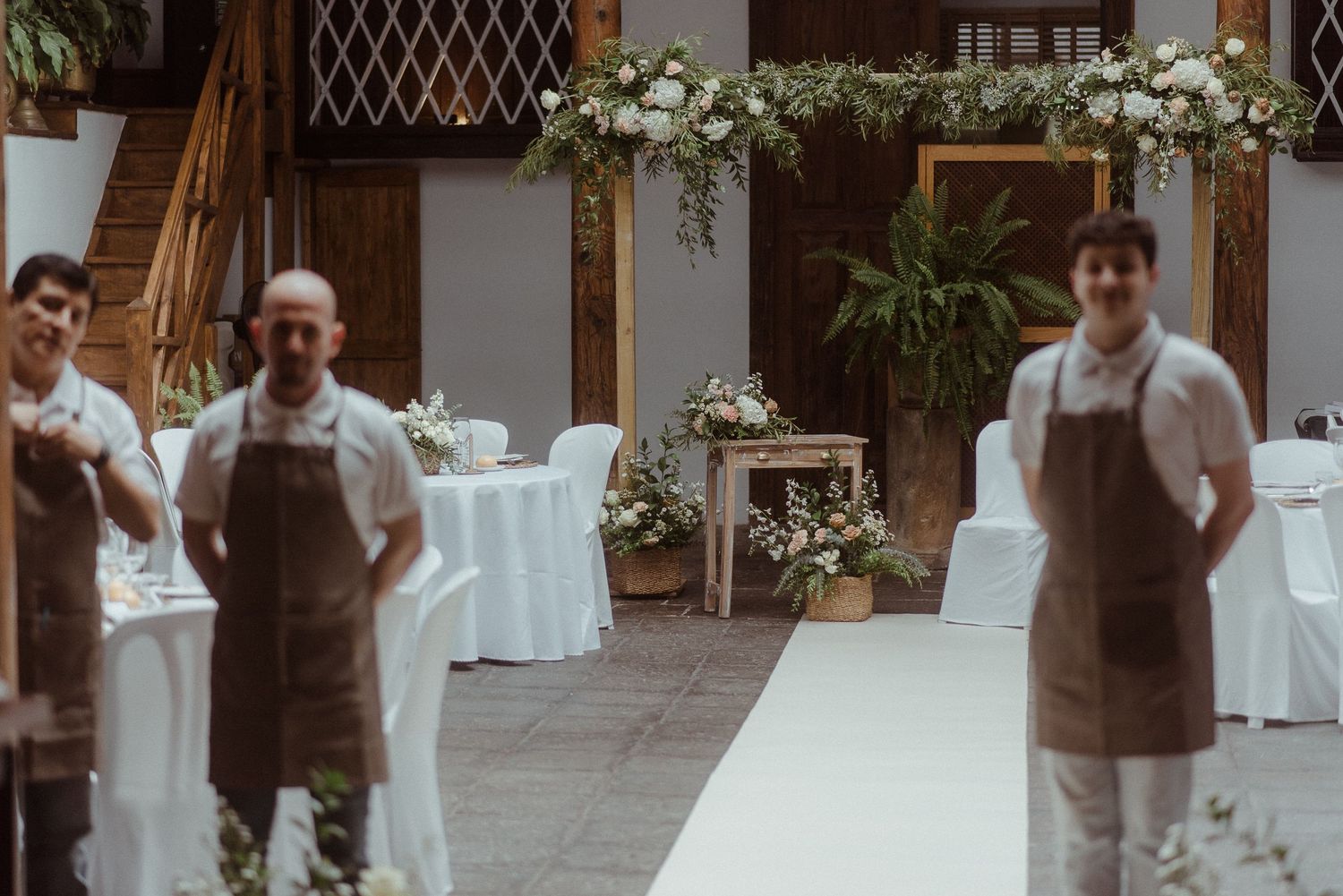 Staff in brown aprons stand in an elegant wedding venue decorated with white linens and floral arrangements.