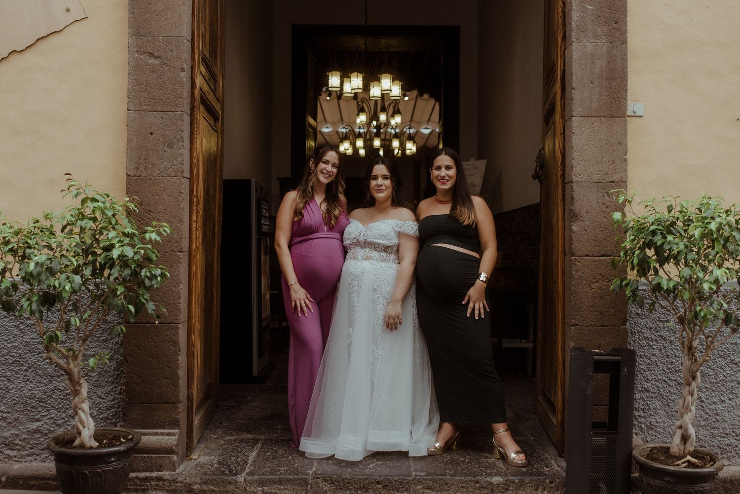 Three people stand together in an elegant doorway with decorative lighting and potted plants on either side.
