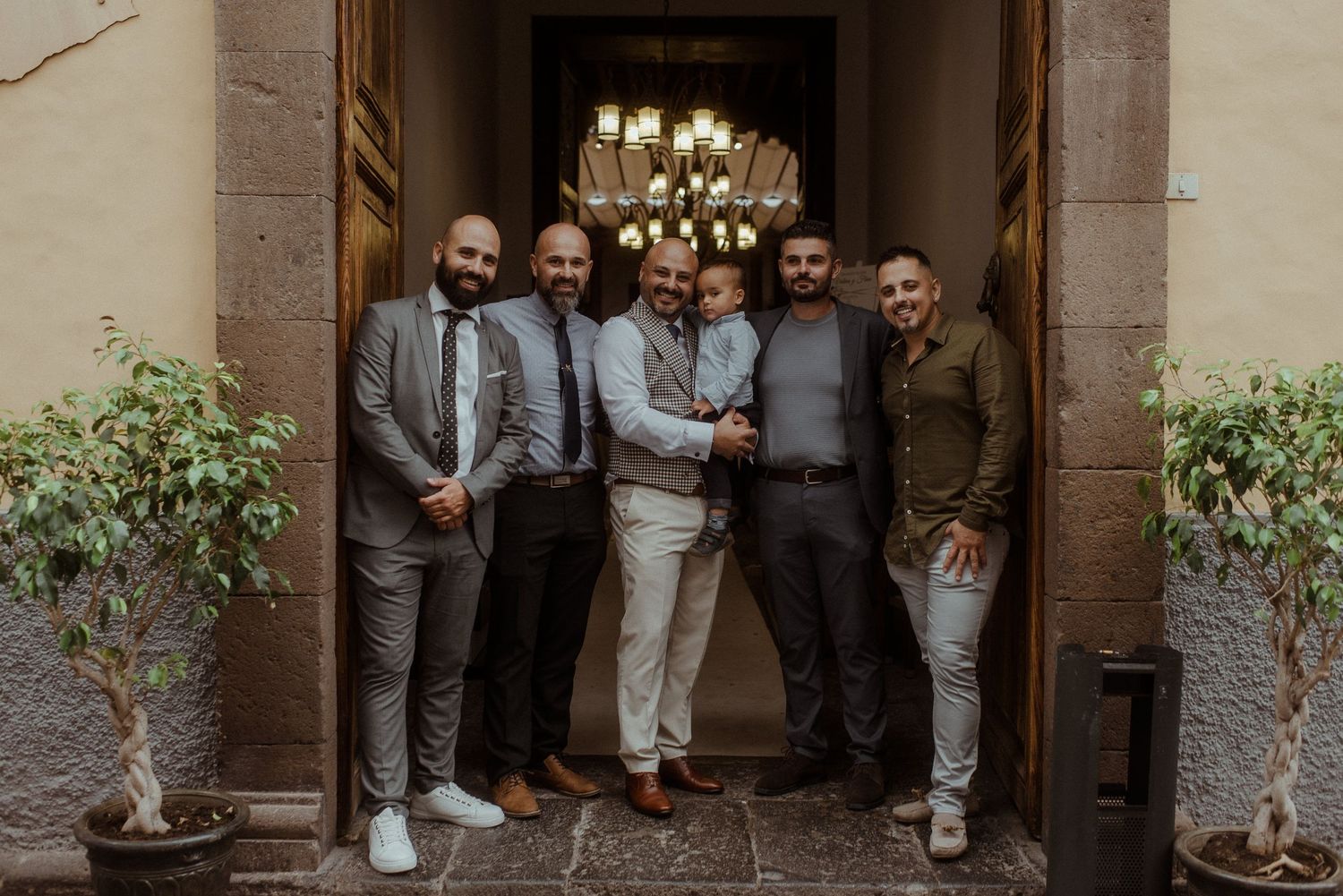 A group of six men in suits and business casual attire stand together in a stone doorway with a chandelier above.