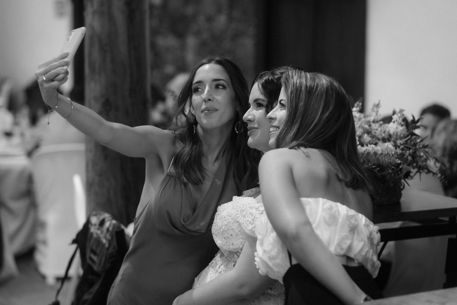Three people take a selfie together at what appears to be a wedding reception in black and white photography.
