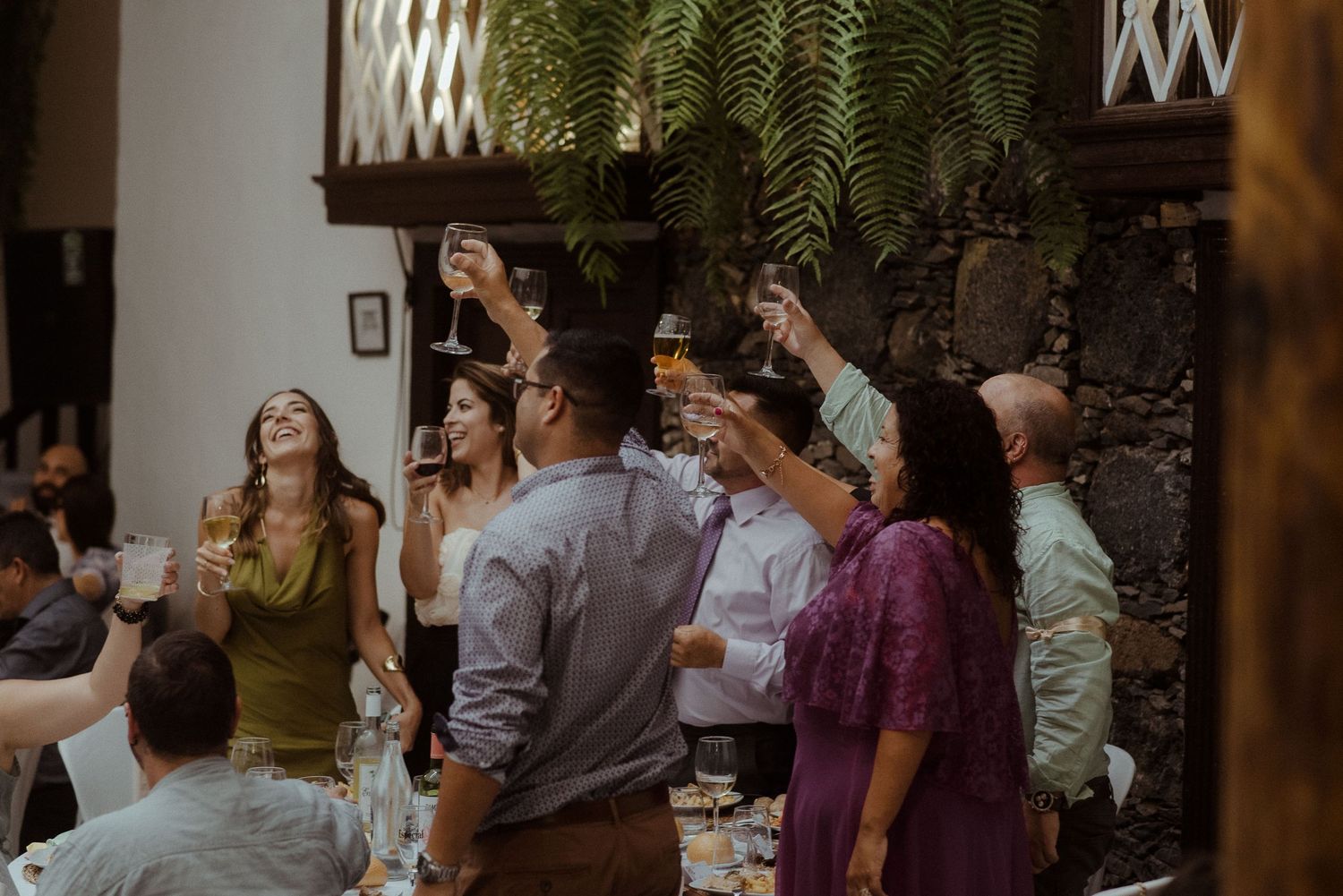 Guests raise their glasses in a toast during an indoor celebration with lush greenery hanging overhead.