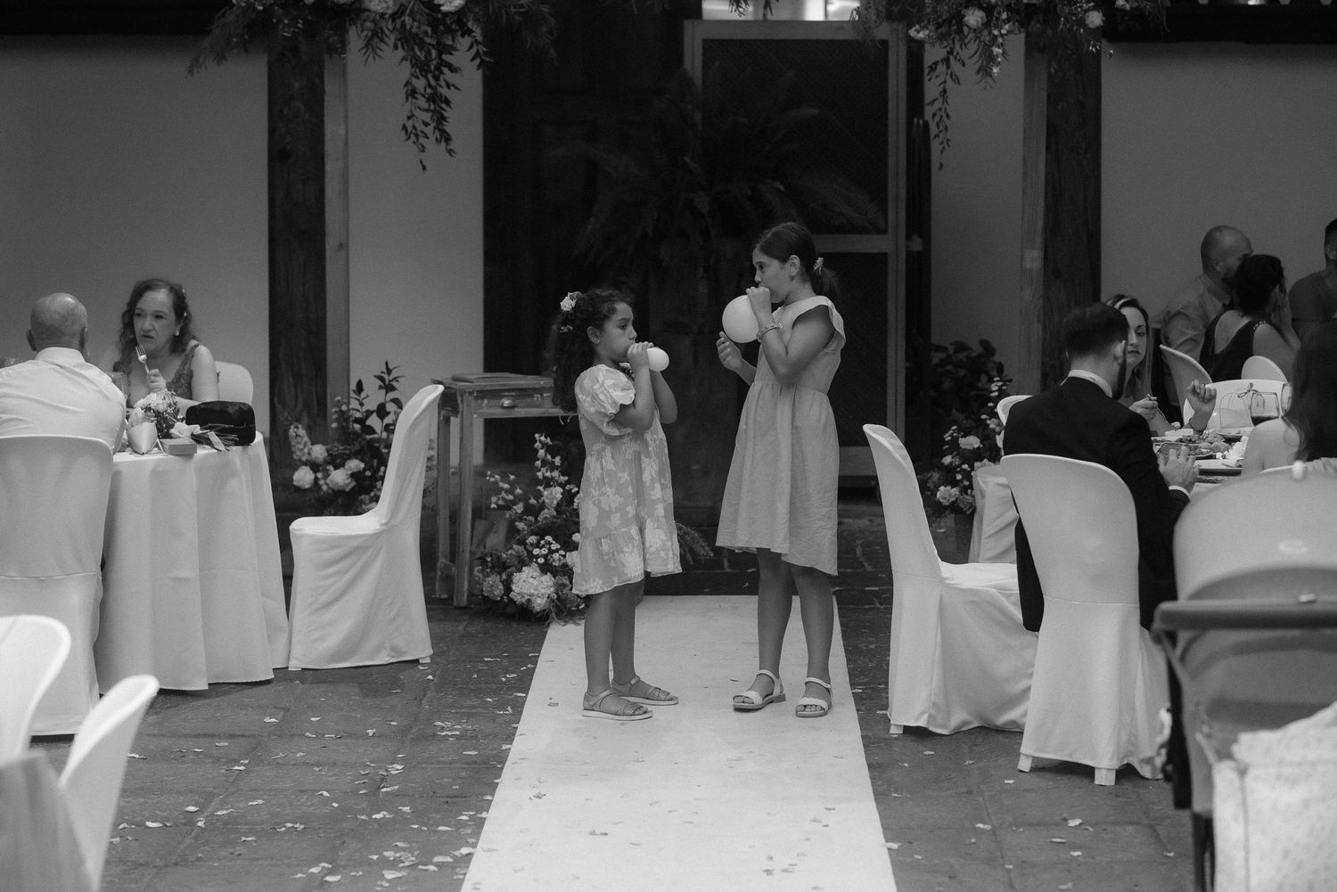 Black and white wedding reception photo showing guests seated at tables as people stand in conversation on a white aisle runner.