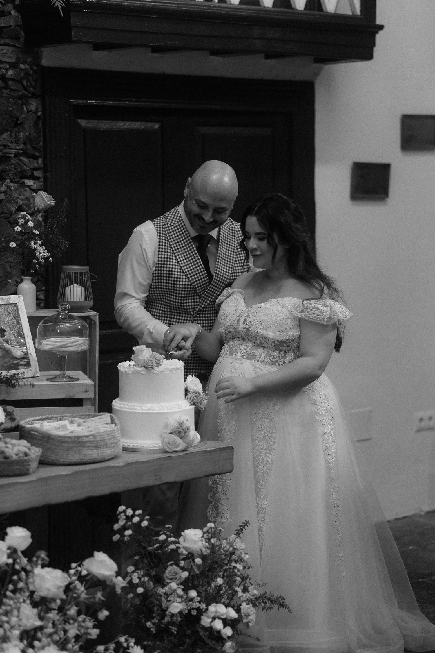 A couple in wedding attire cutting their white two-tier wedding cake together at an outdoor reception with flowers.
