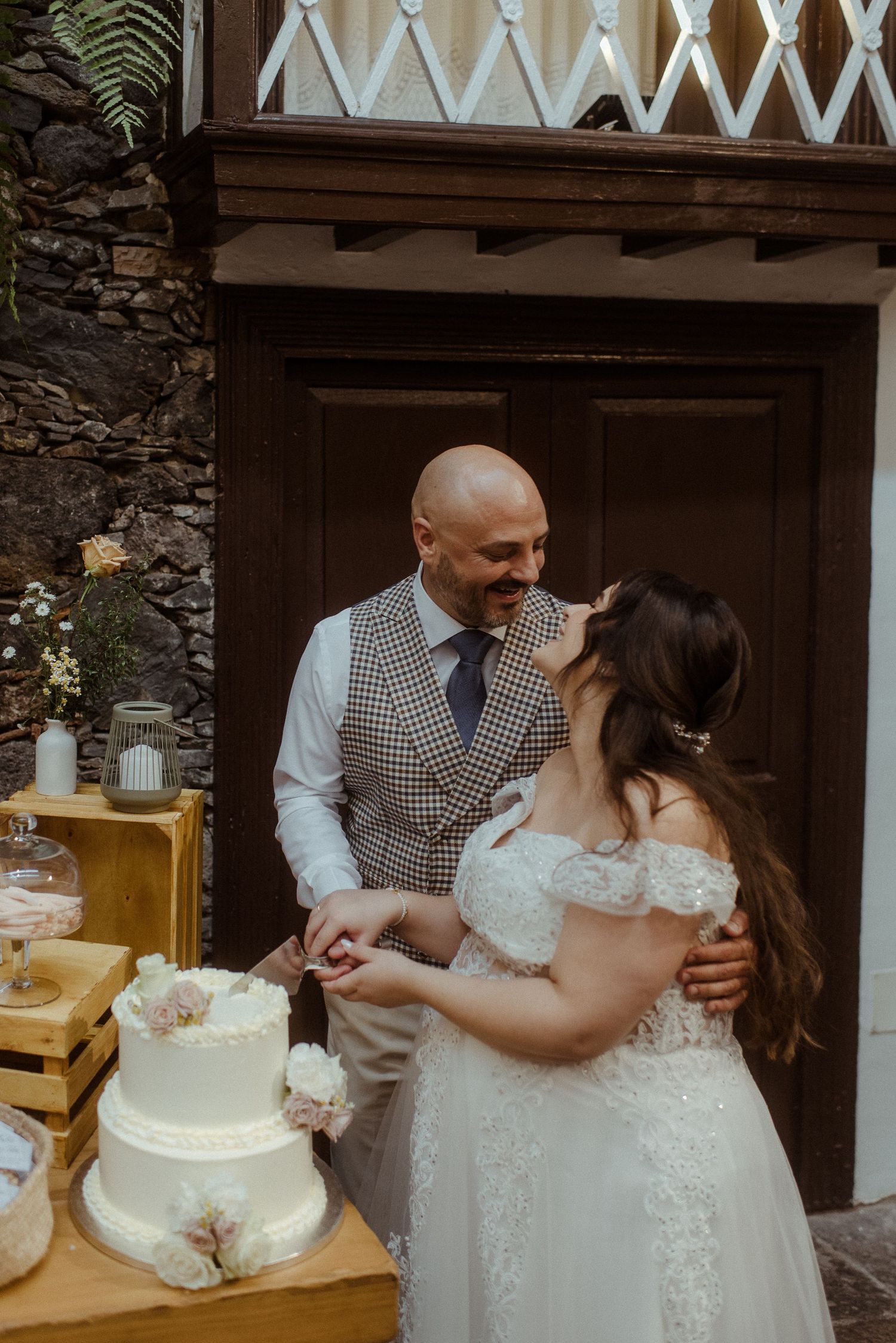 Couple shares intimate moment while cutting their white three-tier wedding cake at rustic outdoor reception.