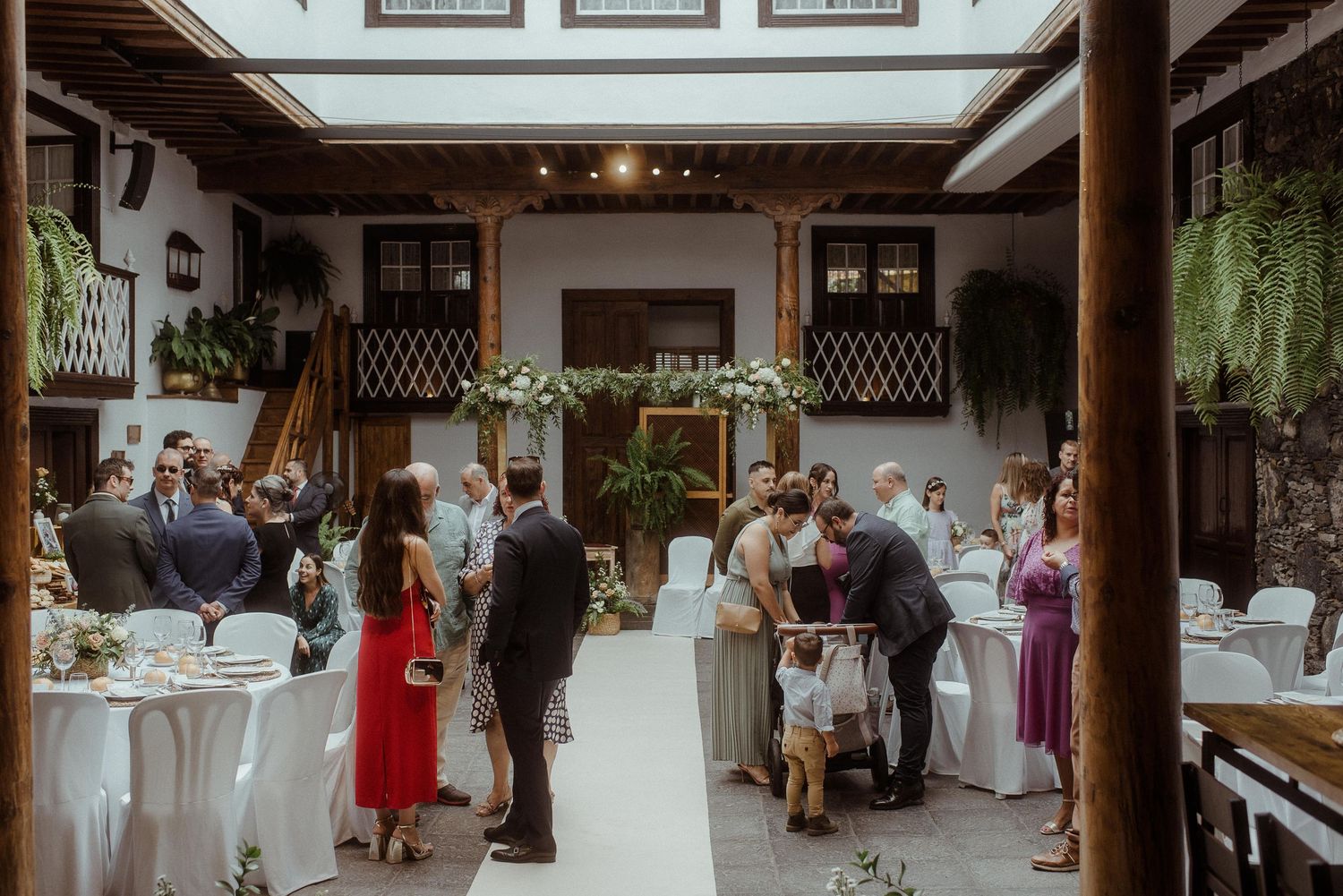 Wedding guests gather at an elegant indoor reception venue with white tables and floral decor.