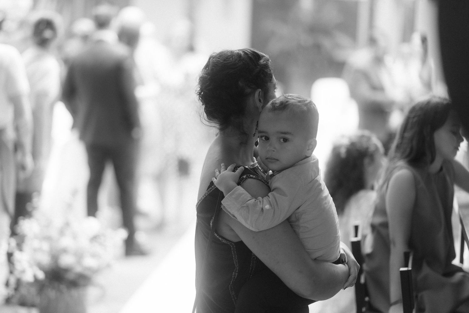 A parent holds a baby at a crowded outdoor gathering in black and white.