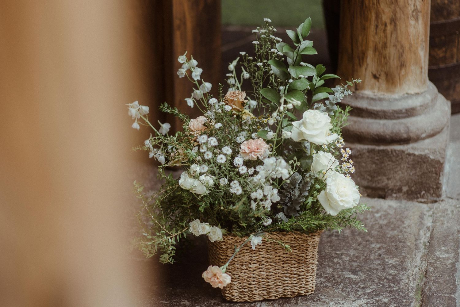 A delicate floral arrangement with white roses and greenery displayed in a rustic wicker basket against stone pillars.