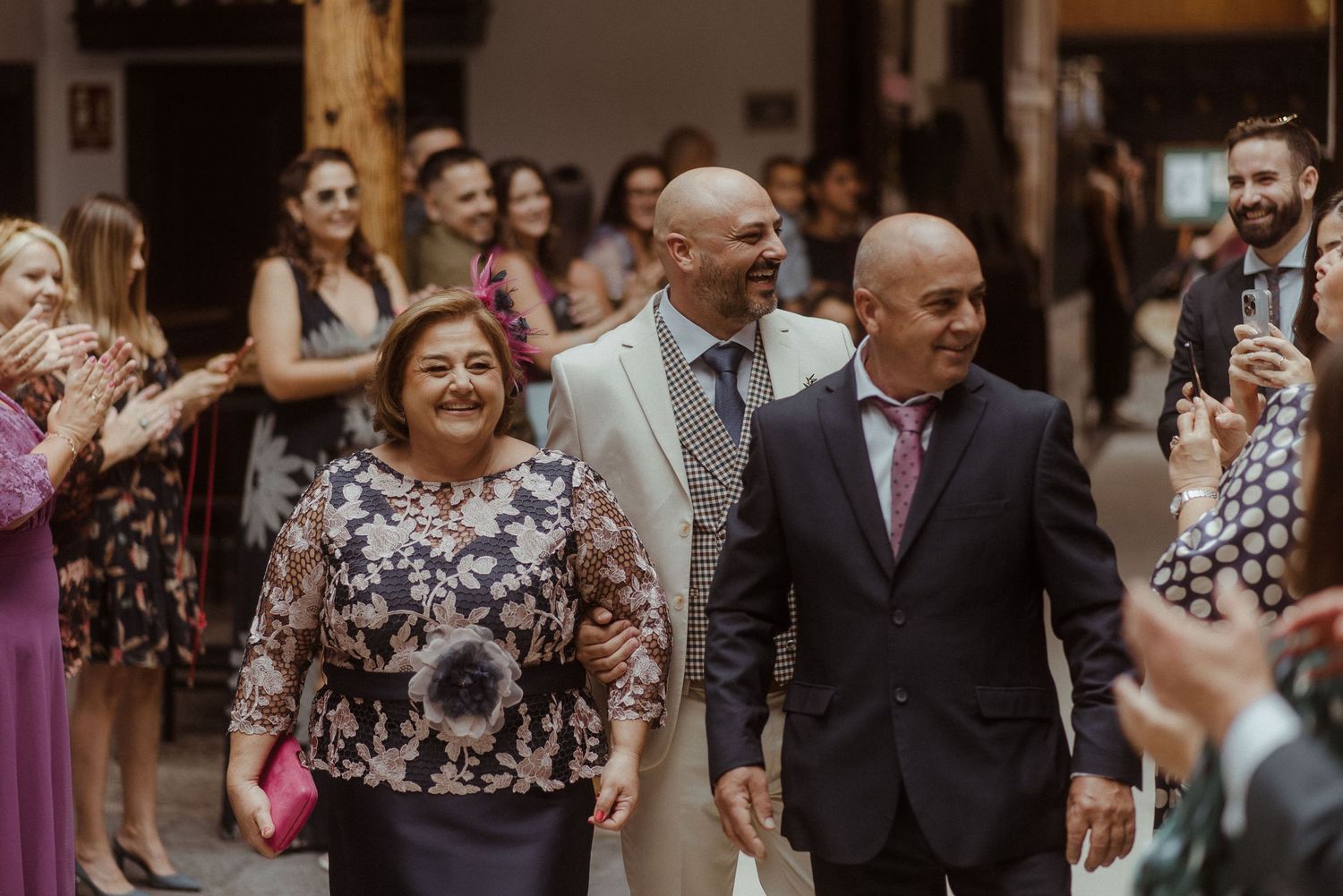 A smiling couple walks down an aisle while wedding guests in colorful attire applaud and celebrate.