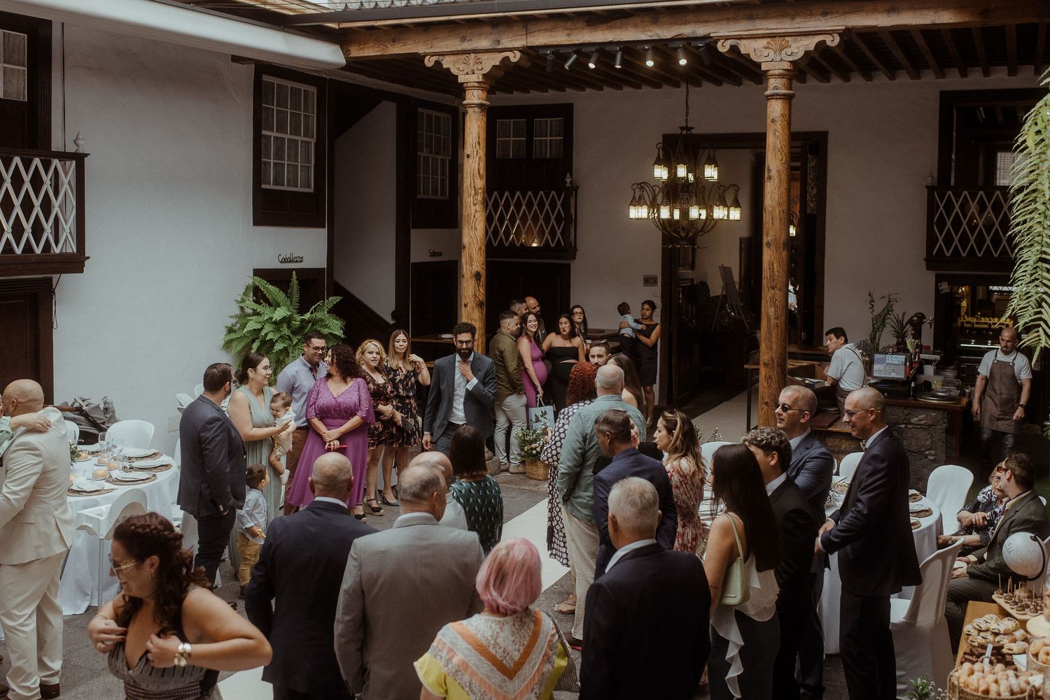 A crowded indoor reception at an elegant venue with wooden columns and chandeliers overhead.