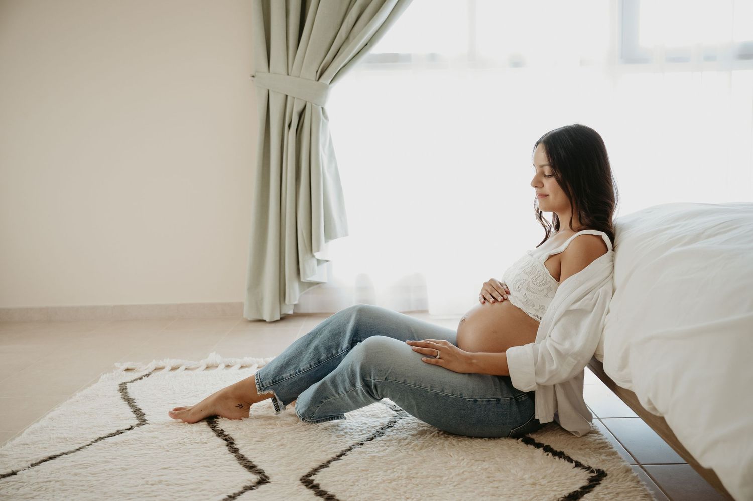 Expecting mother relaxes on geometric rug by window in casual home maternity photo.
