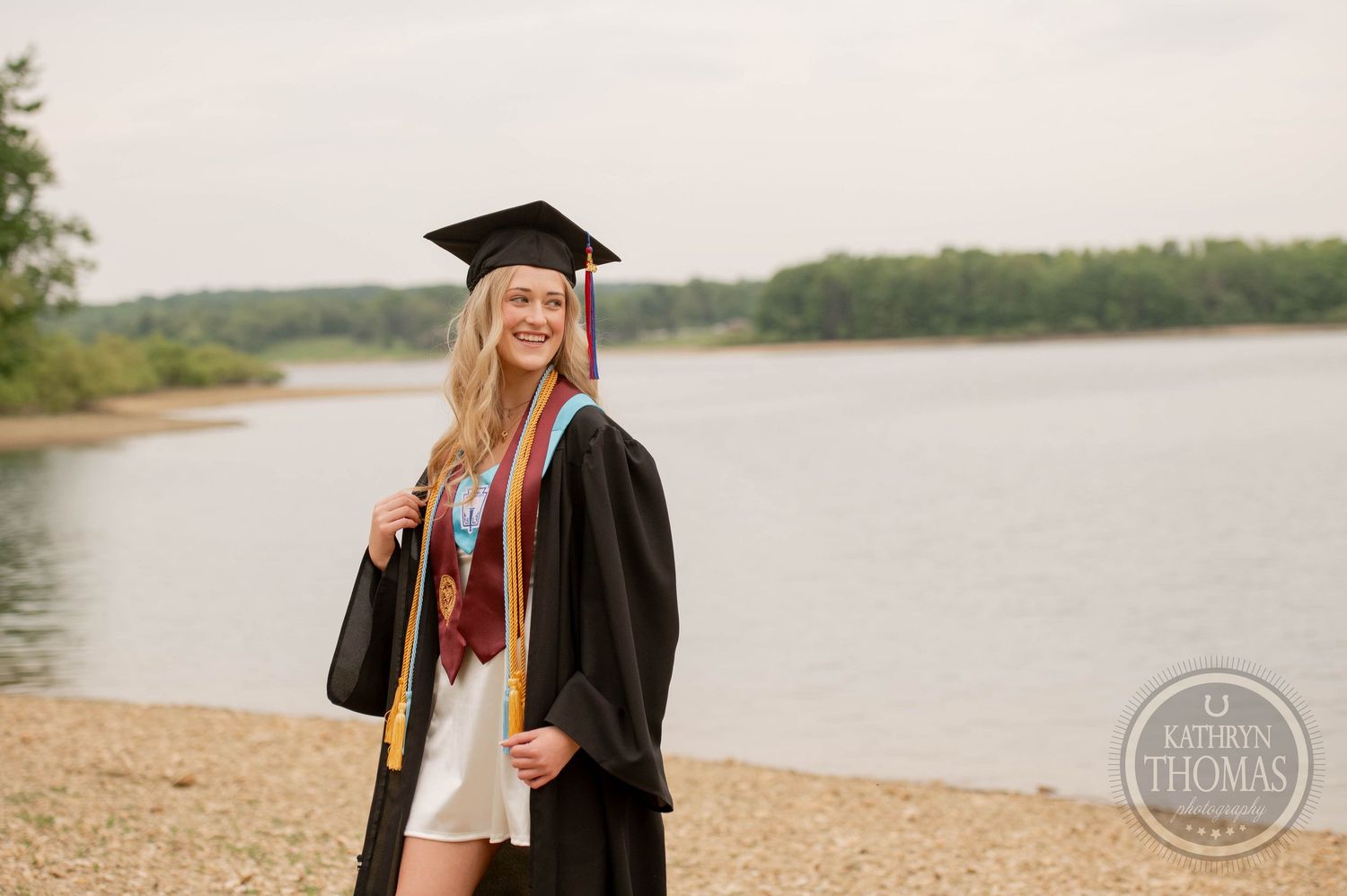 Graduate in cap and gown poses on sandy beach shore with lake and trees in background during graduation photoshoot.