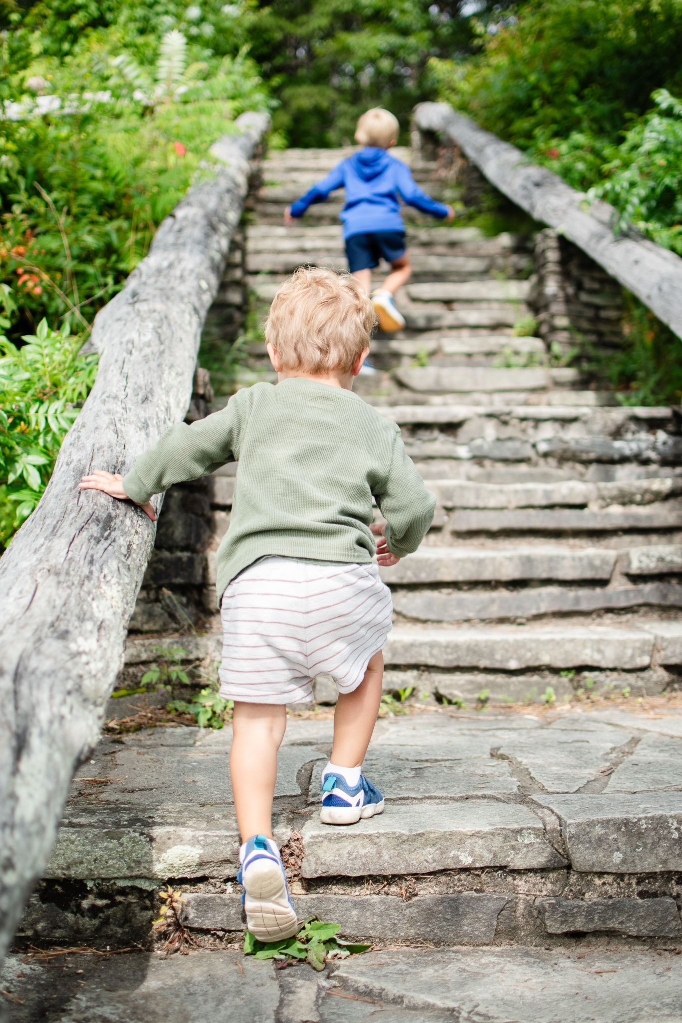 Toddler climbing stone steps in the garden wearing light colored clothing and blue sneakers.