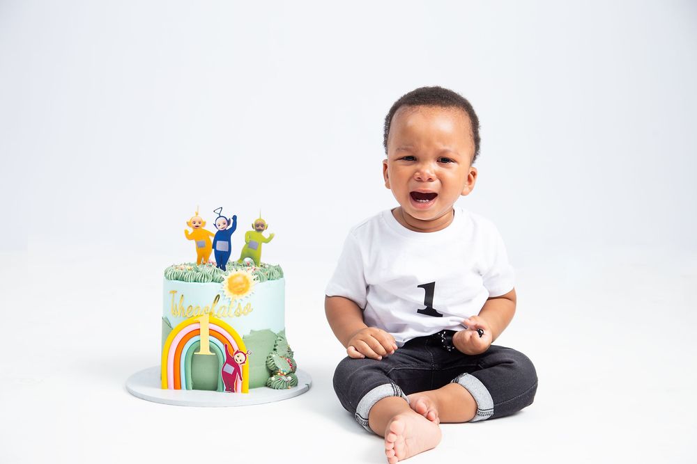 First birthday celebration setup with colorful cake featuring cartoon animals next to smiling child in white shirt.