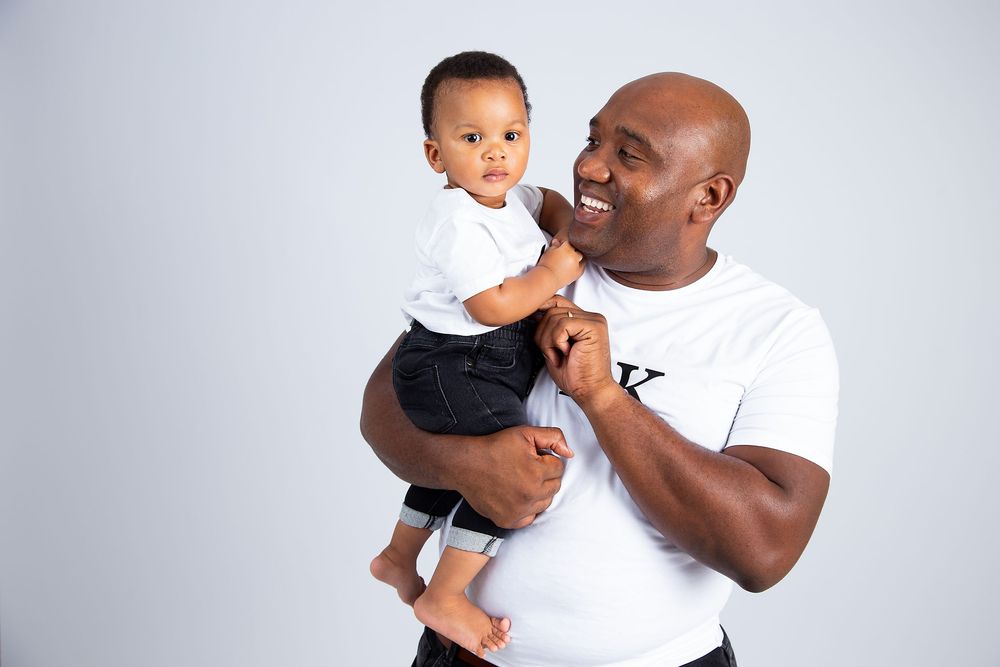 A caring adult holds and smiles at a young child wearing white against a light gray background.
