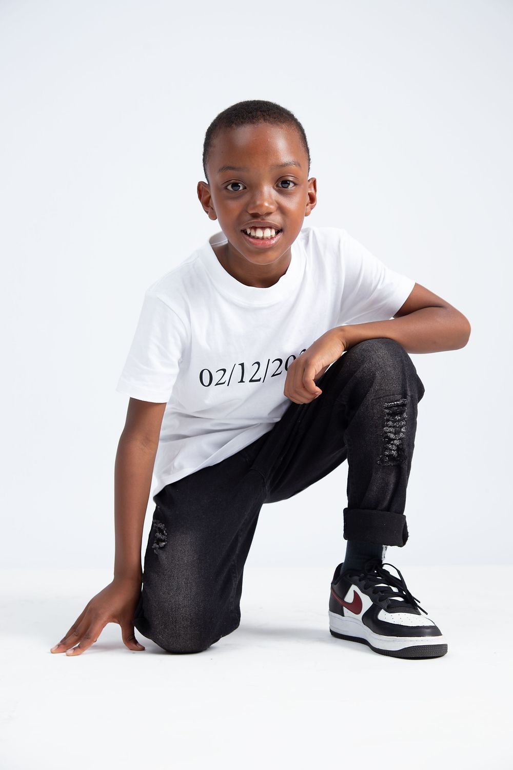 Young person in a white t-shirt with a number date and distressed black jeans kneels while posing against white background.
