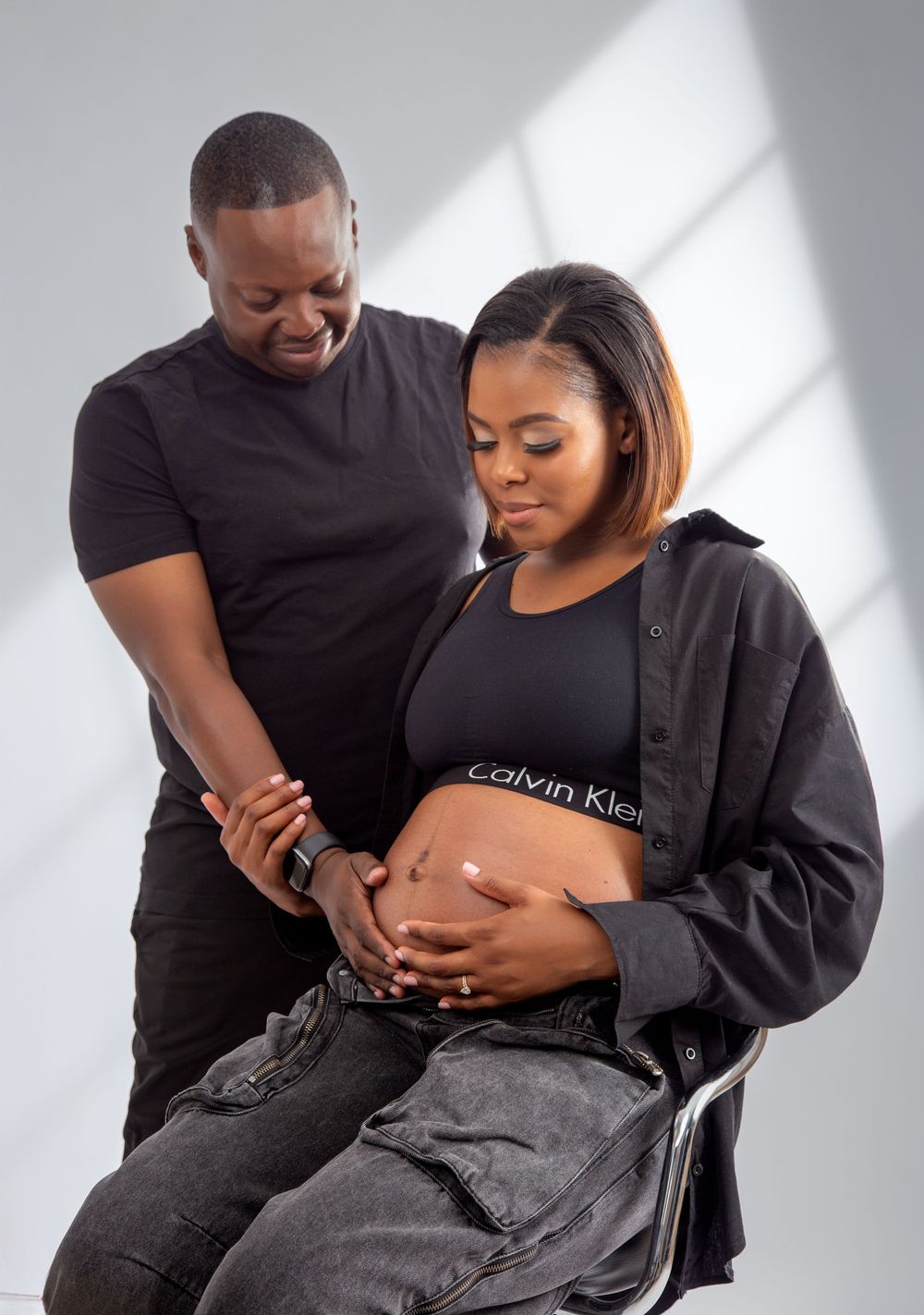 Expecting couple wearing black clothing share intimate moment as they touch pregnant belly in studio with dramatic lighting.
