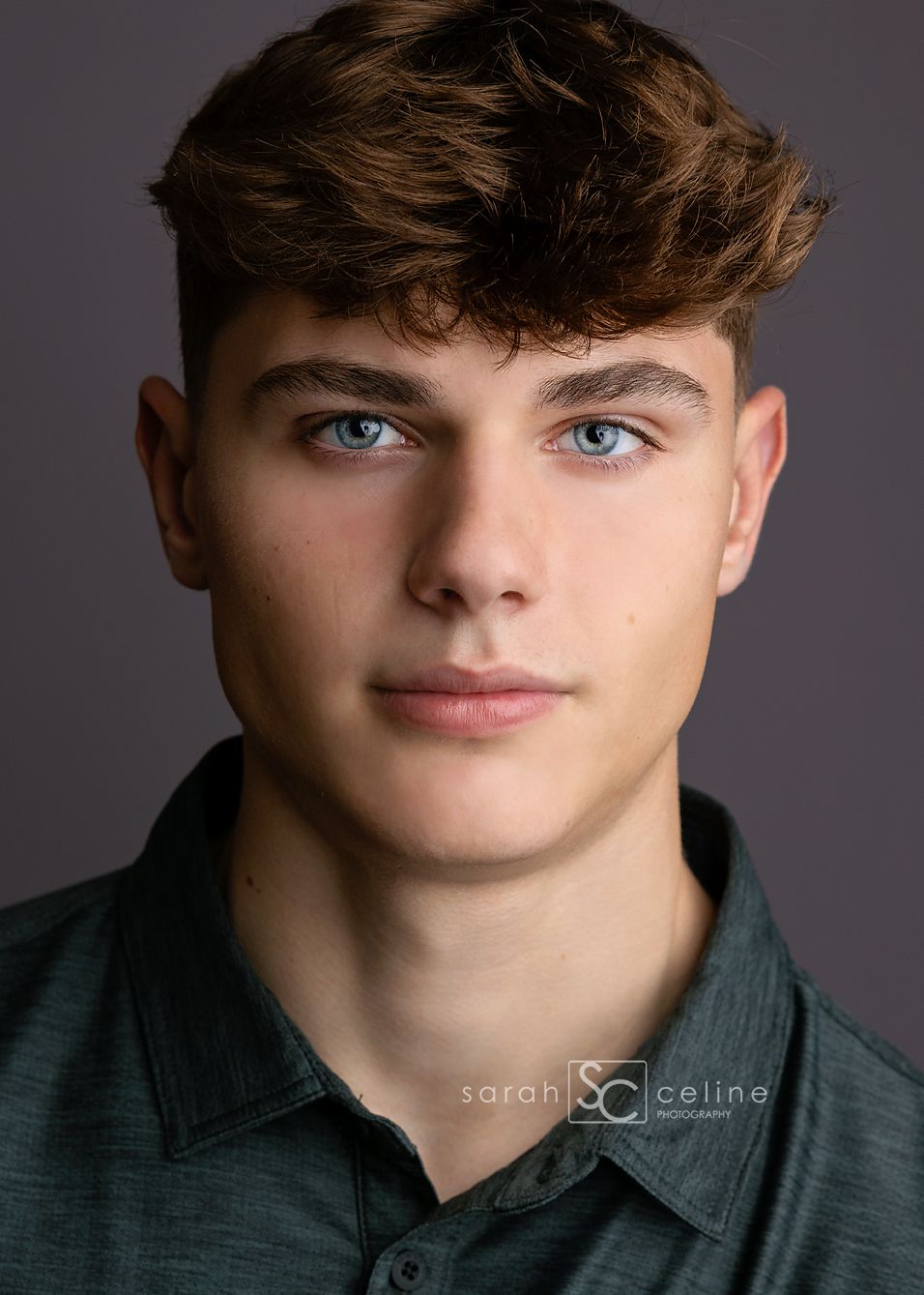 Professional headshot portrait of a young person with wavy brown hair and blue eyes wearing a dark green collared shirt.