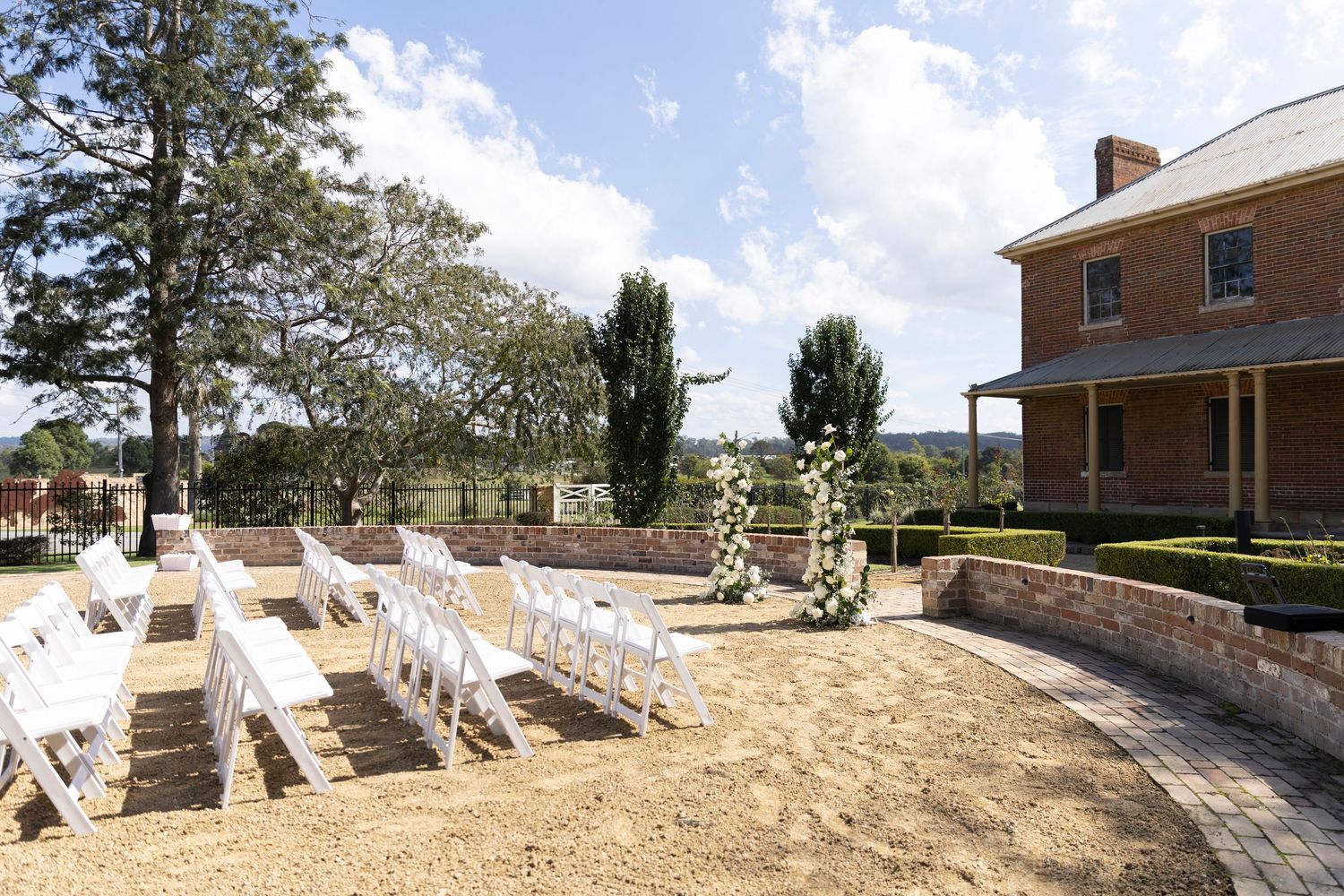 Outdoor wedding ceremony setup with white chairs and benches arranged on sandy ground in front of a historic brick farmhouse.
