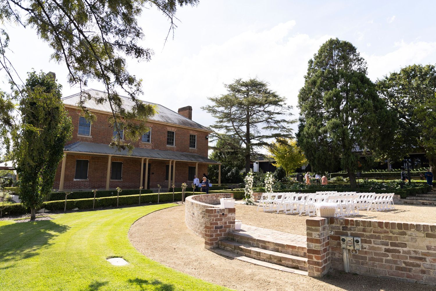 A stone wall leads up a grassy hillside path to a brick building on a sunny day.