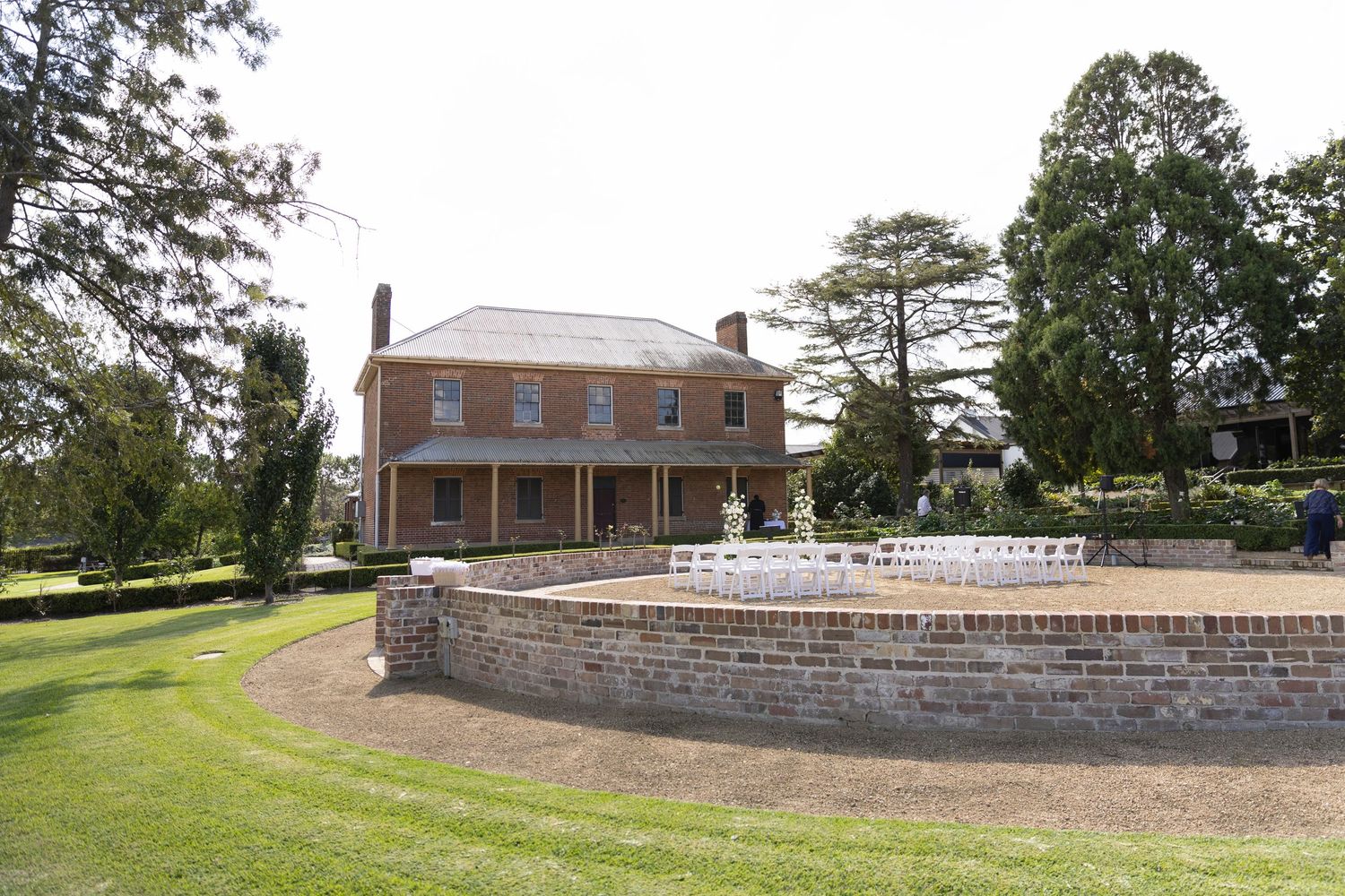 Historic brick mansion with curved stone retaining wall surrounded by manicured lawn and tall trees on sunny day.