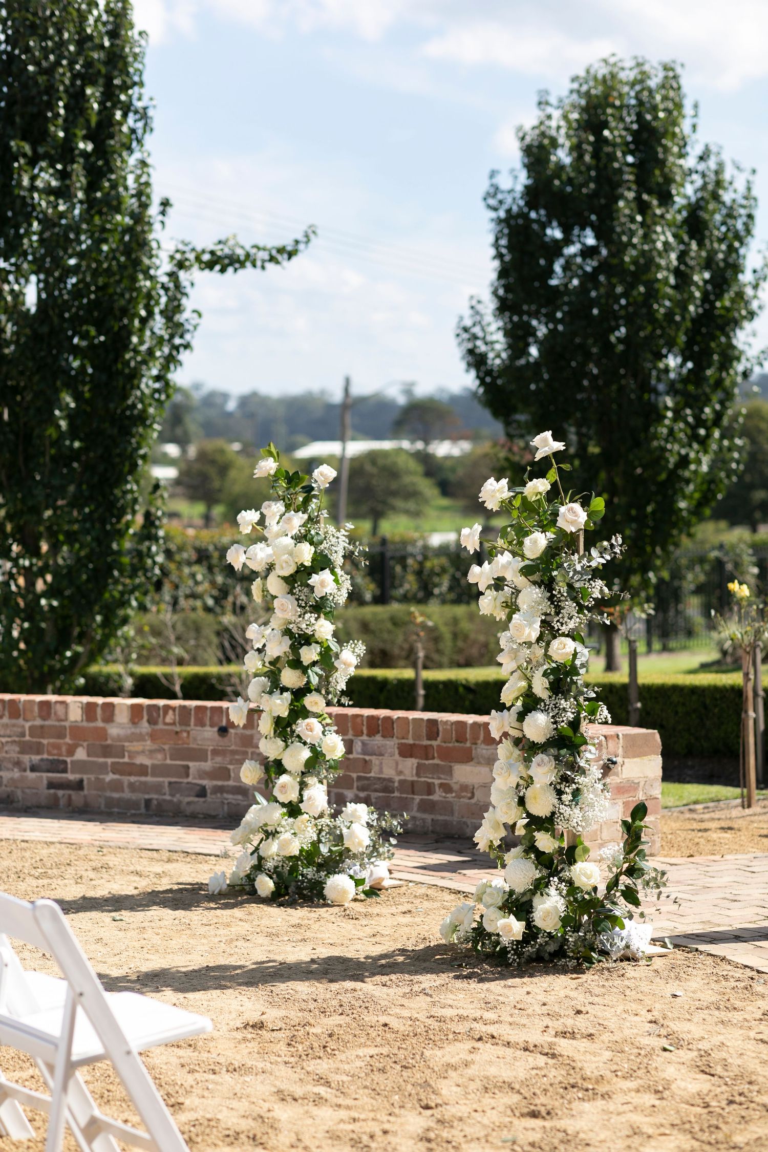 White floral wedding arch with roses stands against a brick wall in an outdoor garden setting.