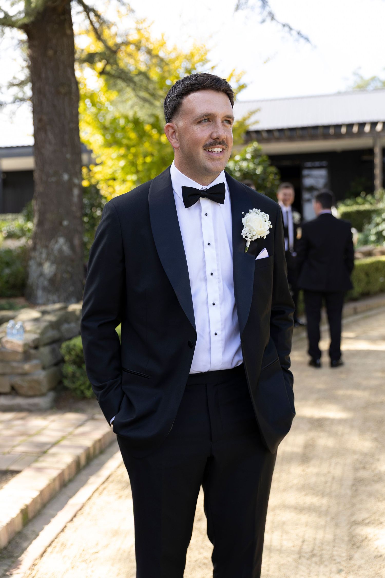 A groom in a classic black tuxedo and bowtie smiles during an outdoor wedding.