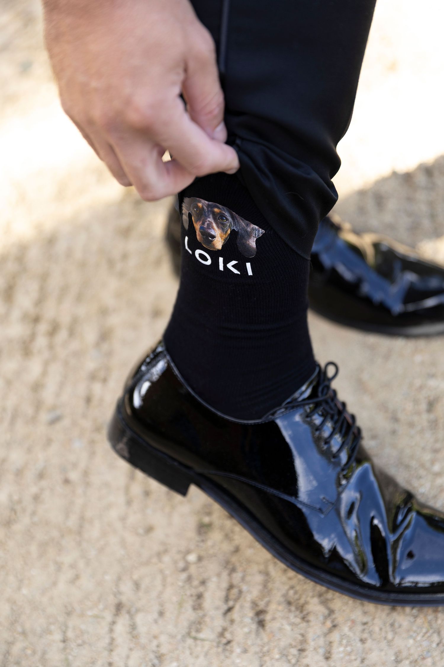 A person wearing shiny black patent leather dress shoes and dark socks with 'LOKI' branding pulls up their sock.