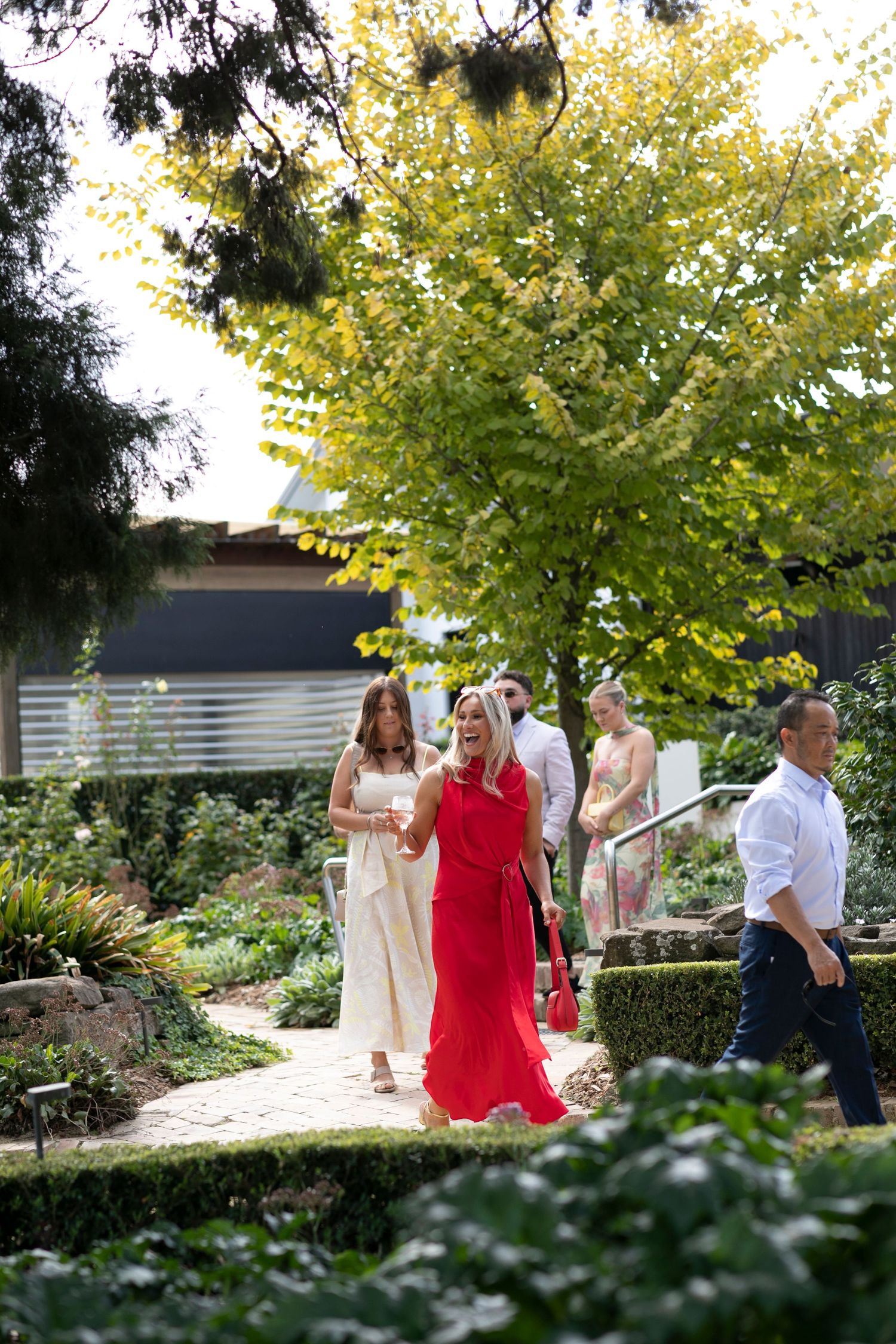 Wedding guests gather under a blooming yellow tree in a garden pathway during an outdoor celebration.