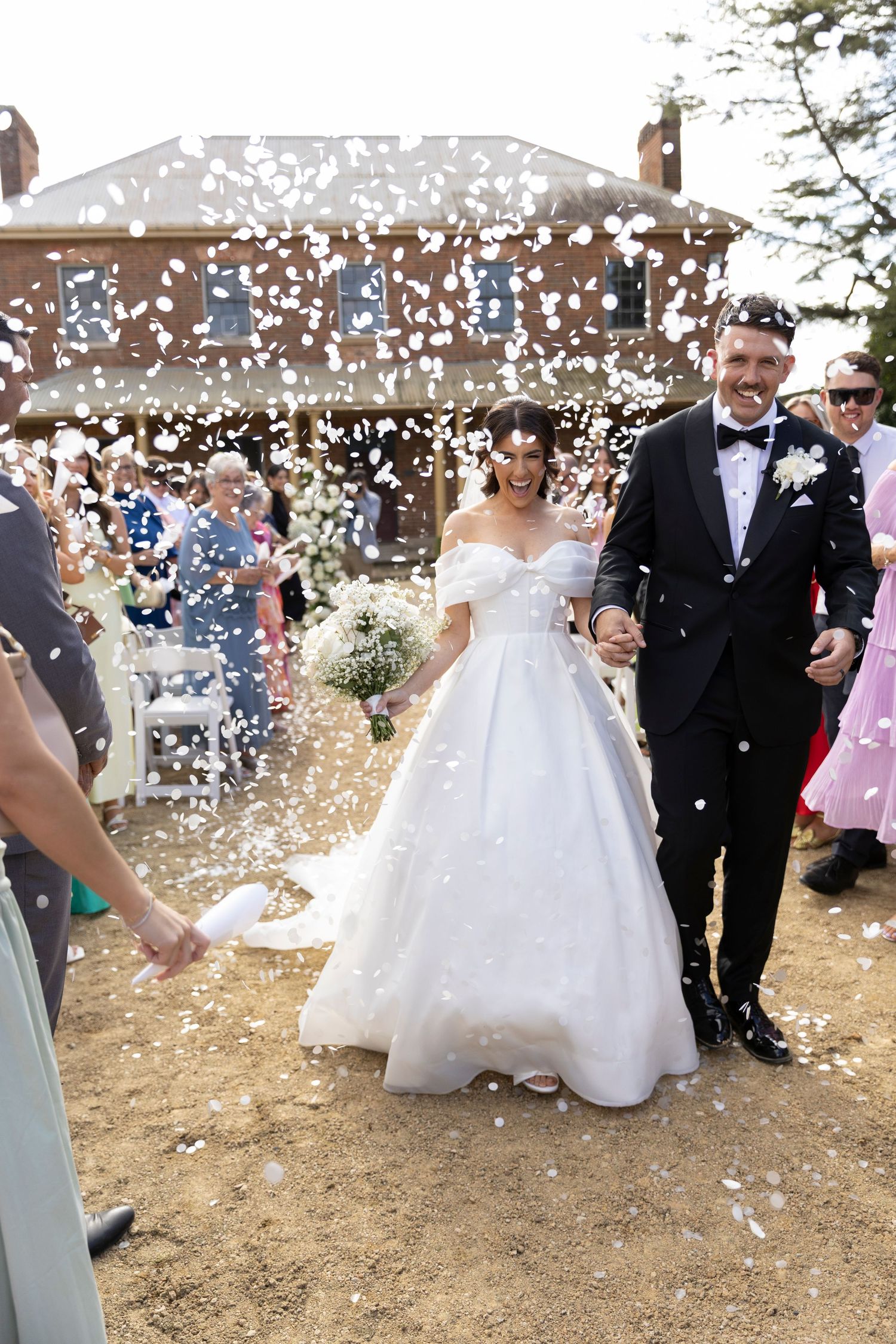 Wedding couple beams with joy while walking through white confetti outside historic brick venue.