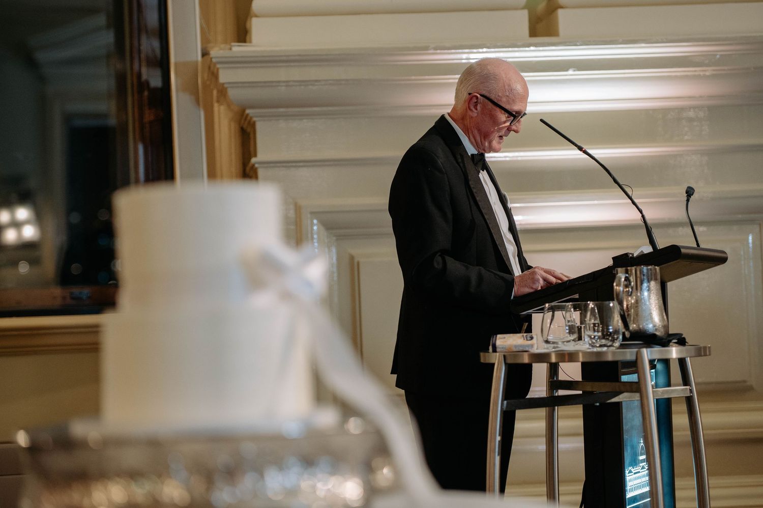 A speaker stands at a podium in a formal event setting with warm lighting and elegant decor.